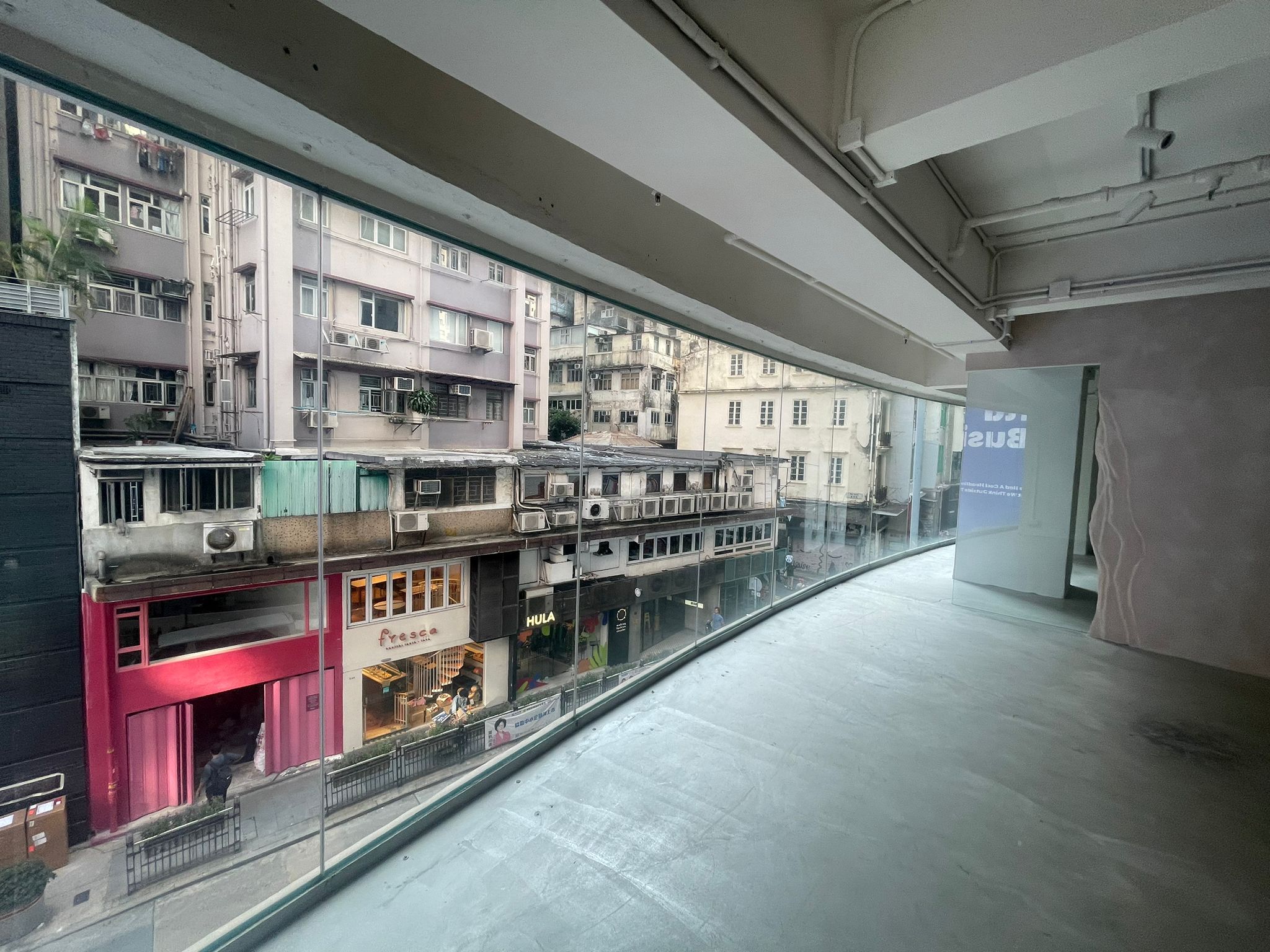 View from Ref#2980, an empty modern space with large curved glass windows ideal for office rental overlooking a city street lined with older, densely packed buildings and shops.