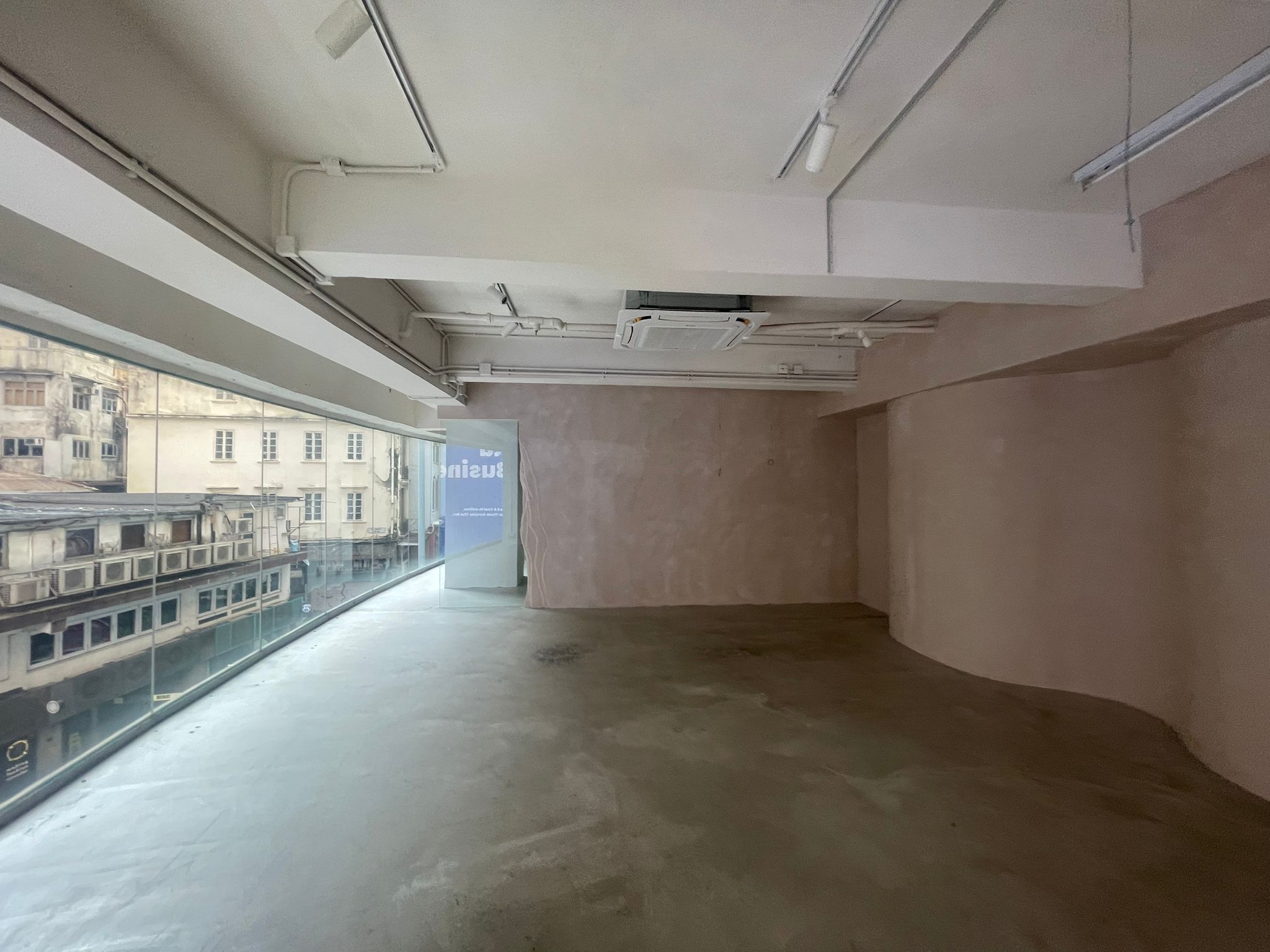 Ref#2980 features an empty room with bare walls, concrete floor, and a large curved window overlooking old buildings. Well-lit with ceiling lights and visible air conditioning ducts, this space is ideal to rent as an office or shop.