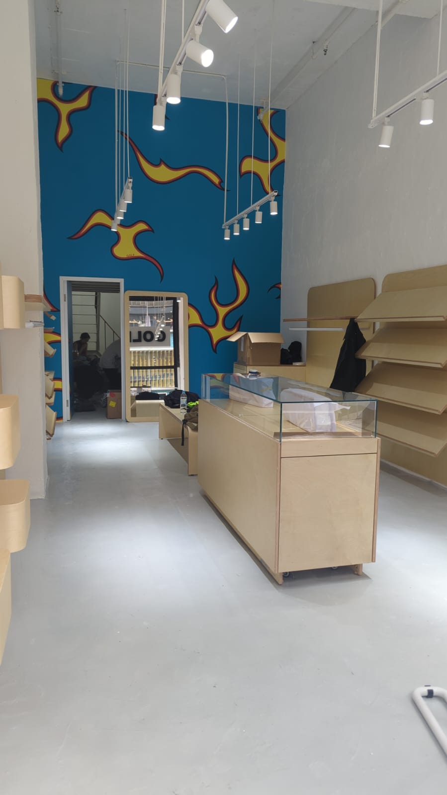 Ref#2985: Modern retail space for rent with empty wood shelves, glass display cases, white floors, track lighting, and a bold blue mural with yellow and orange flames—ideal to rent for a shop or restaurant.