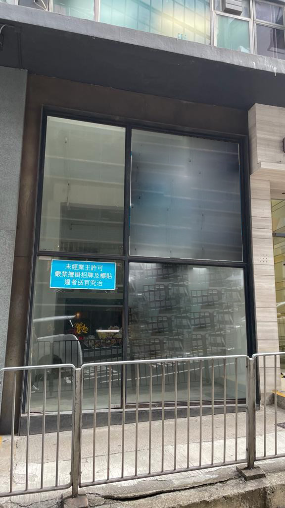 A large glass window displays a blue Chinese sign, likely for rent, on the modern Ref#2985 building. Metal railings line the sidewalk, while reflections and interior shelves are visible inside.