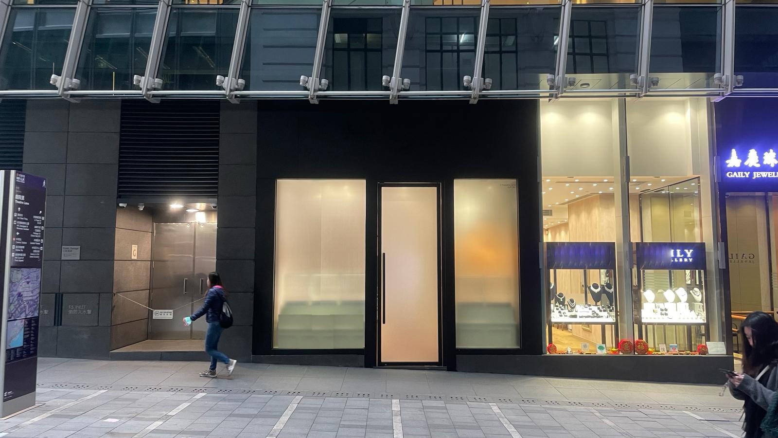 A person passes Ref#3046, a modern building with a large frosted glass door, perfect to rent as an office or shop. Beside it is a brightly lit jewelry store with illuminated displays, highlighting the quiet street.