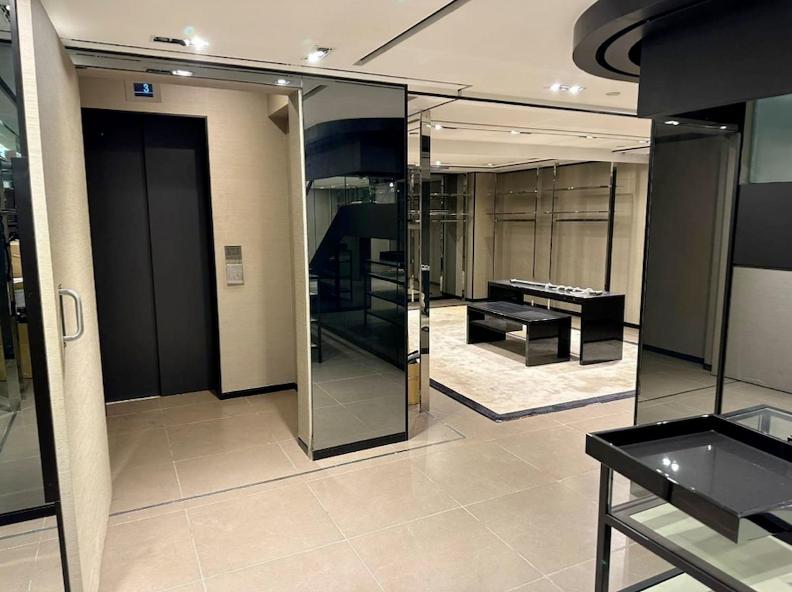 Ref#3120: A modern, empty retail store with tiled floors, glass display cases, a beige rug, black tables, and an elevator on the left. Well-lit with recessed ceiling lightsideal to rent shop or start your dream business.