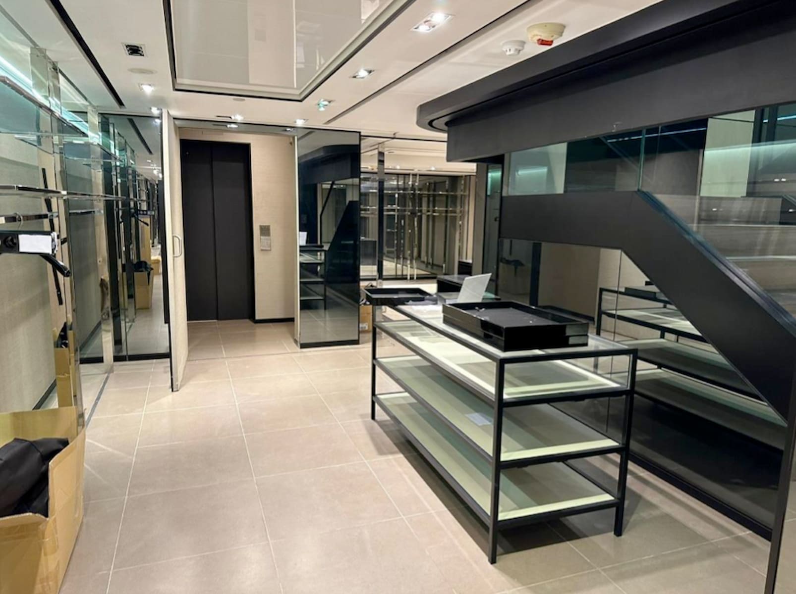 Ref#3120: A modern, empty retail space with tiled floors, glass walls, shelves, and a sleek black staircase. Boxes on the floor and a laptop on one shelf show an organized but unfinished store interior ready to rent.