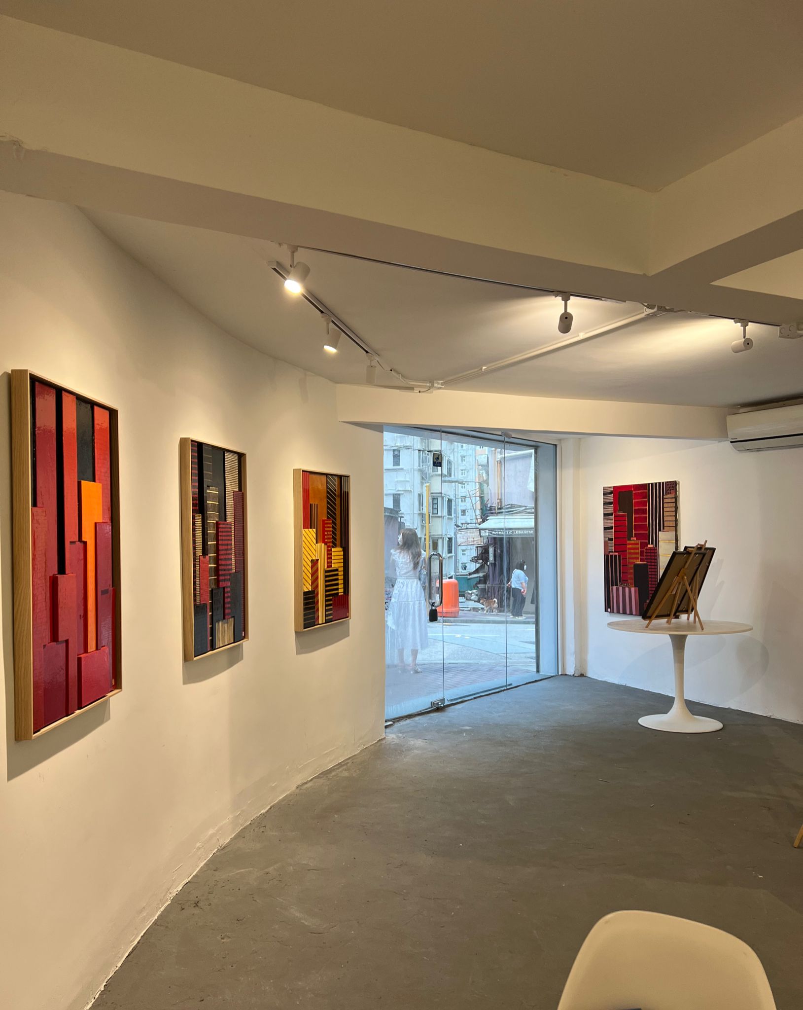 Ref#2988: A modern art gallery with white walls features four abstract geometric paintings in red, yellow, and brown. Large windows overlook the city—ideal for a stylish office rental. A round white table displays additional artworks.