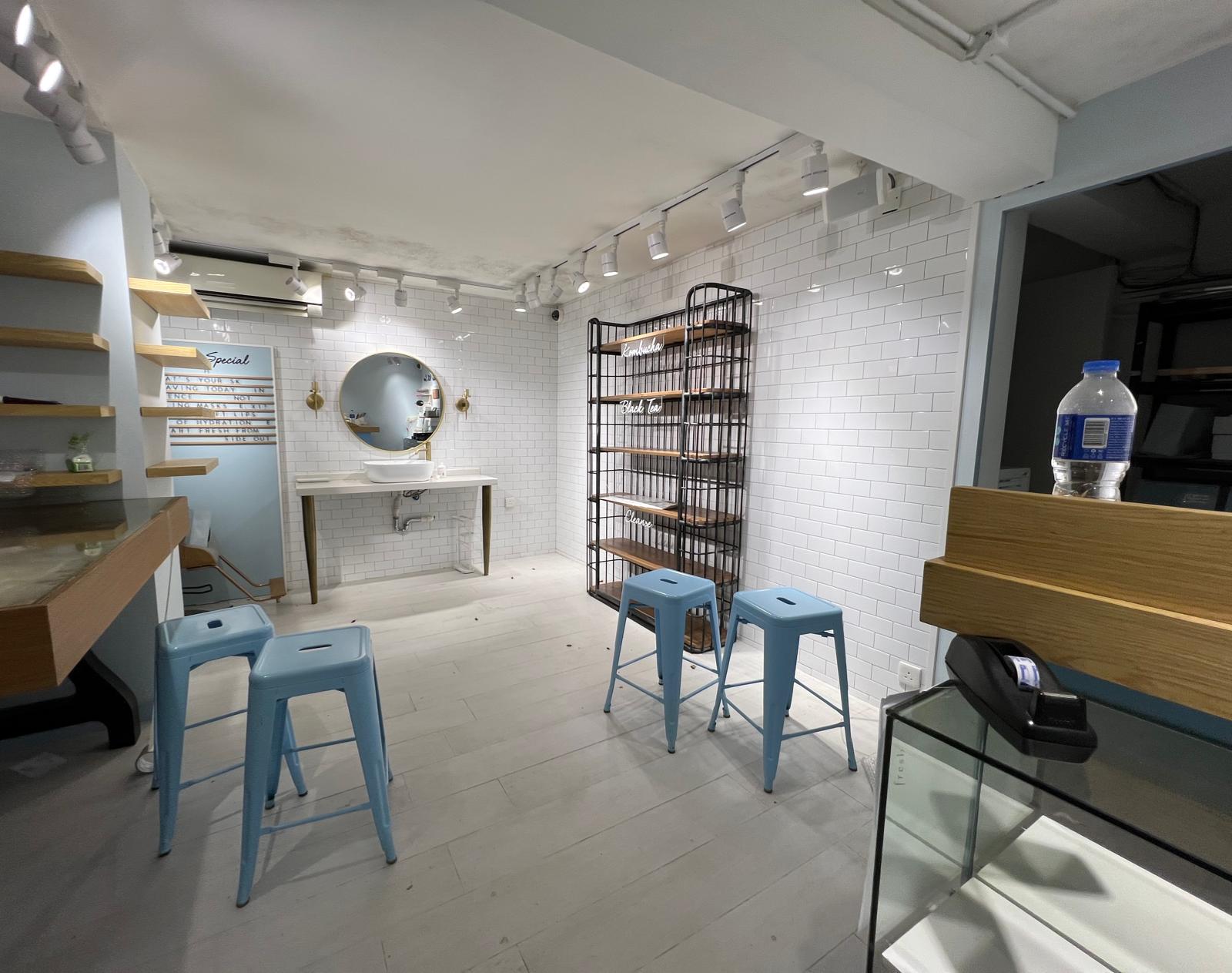 Ref#2988 is a minimalist, modern space ideal for a shop or office, with white subway tile walls, light wood floors, blue stools, empty display shelves, a round mirror above the sink, and bright even lighting.