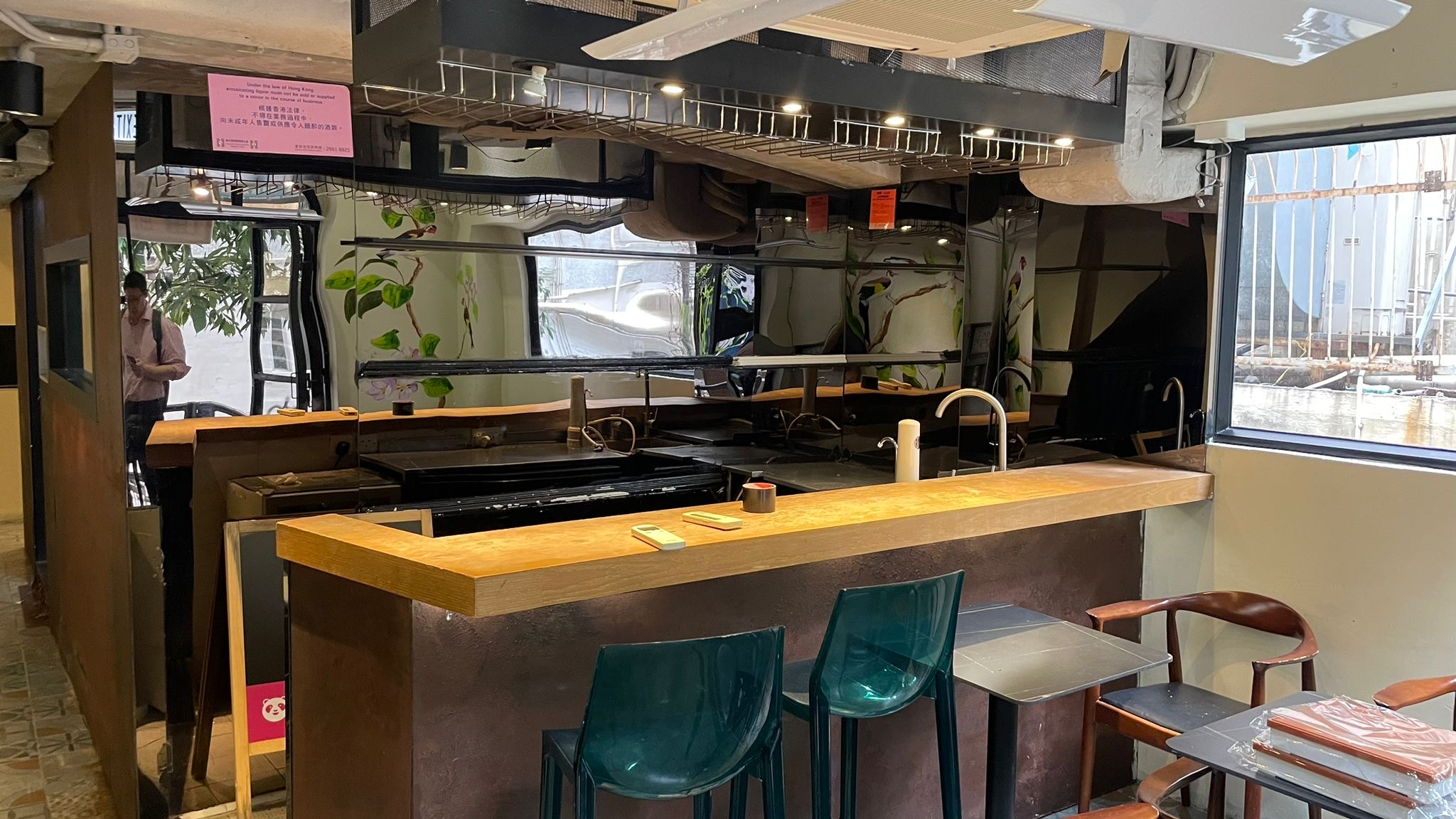 Ref#2994: Modern space ideal for a restaurant, featuring a wooden counter, teal bar stools, brown chairs, large windows filling the bright interior with light, plant-filled walls, and sleek glass reflections.