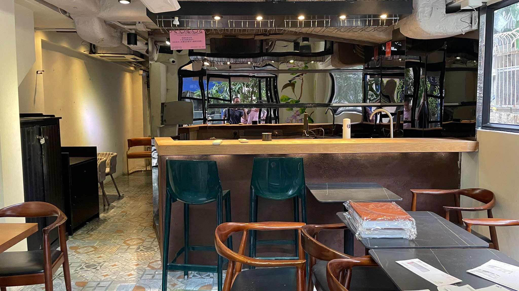 Ref#2994: Inviting modern cafe interior ideal for restaurant rental, with a wooden counter, green barstools, cozy tables with stacked menus and papers. A mirrored wall reflects lush plants and outdoor scenery in this empty space.