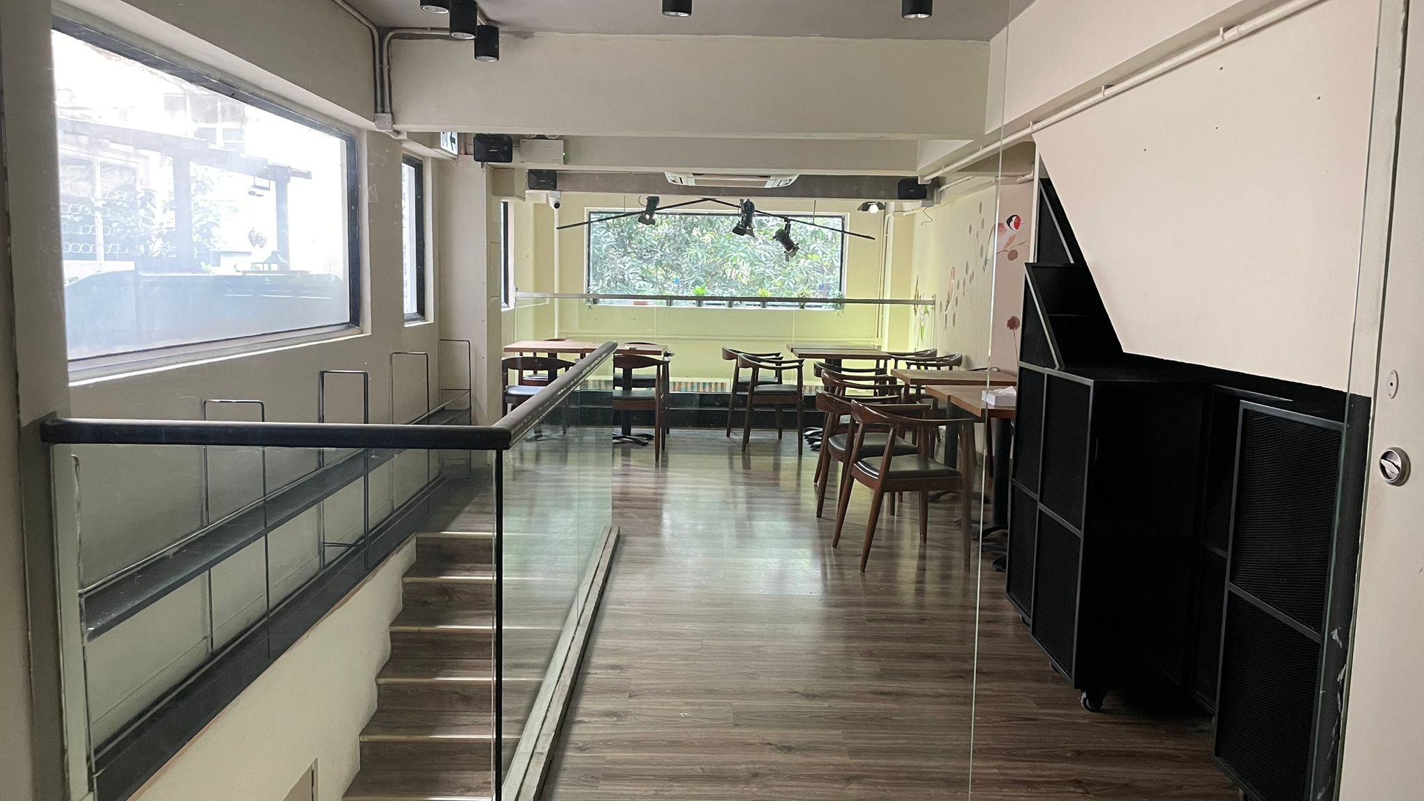 Ref#2994: A modern, empty cafe with wooden floors, tables, and chairs. Large windows brighten the space with natural light; glass-railed stairs lead to seating. Trees are visible through the windows. Ideal for shop or restaurant rental.