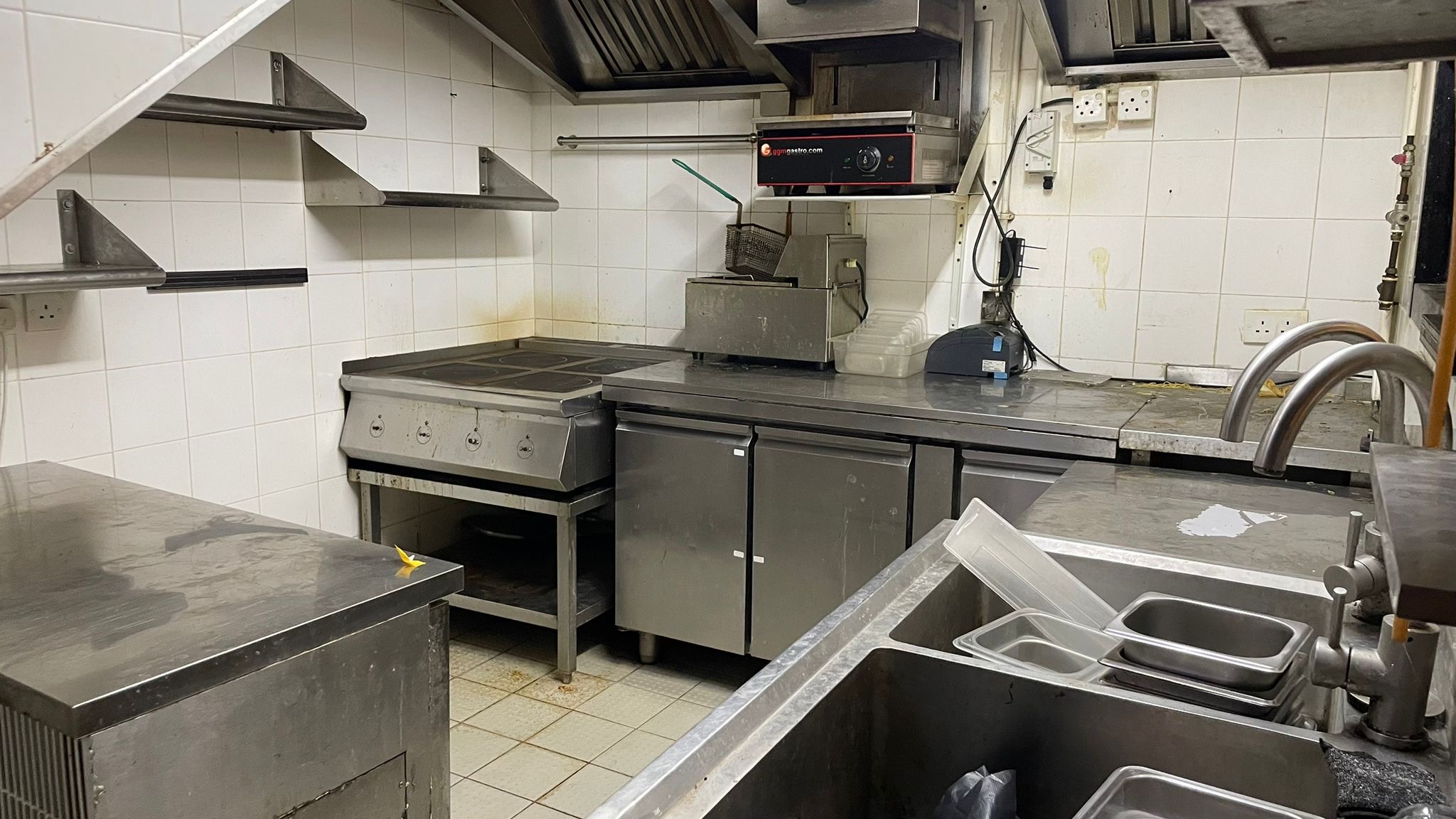 Ref#2994: Commercial kitchen with stainless steel counters, electric stove, deep fryer, metal shelves, sinks, and storage units—perfect for restaurant rental. White-tiled walls emphasize its clean, unused look.