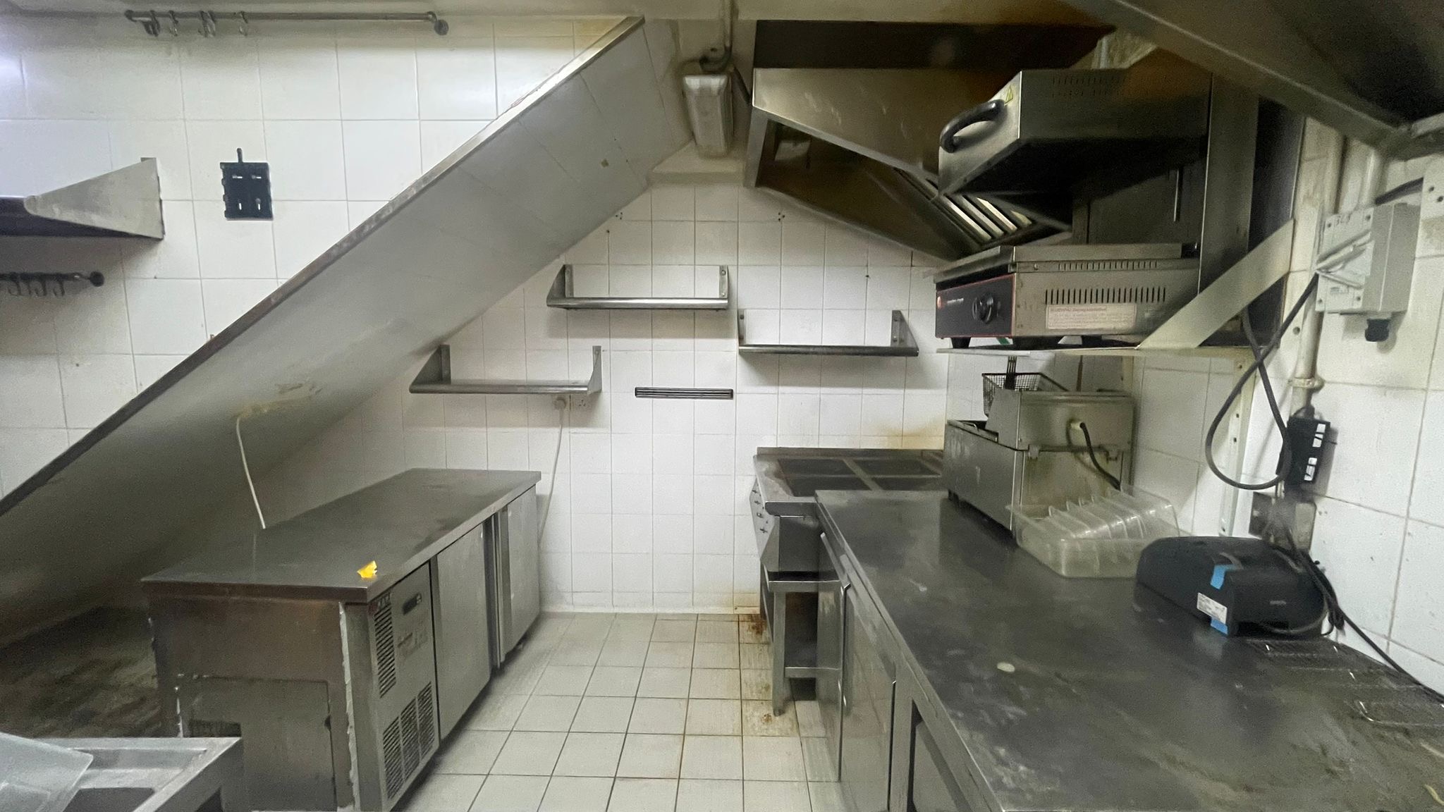 Ref#2994: A commercial kitchen for rent, ideal for a shop or restaurant, with stainless steel counters and appliances, metal shelving, white tiled walls and floors, slanted ceiling, and ventilation hood.