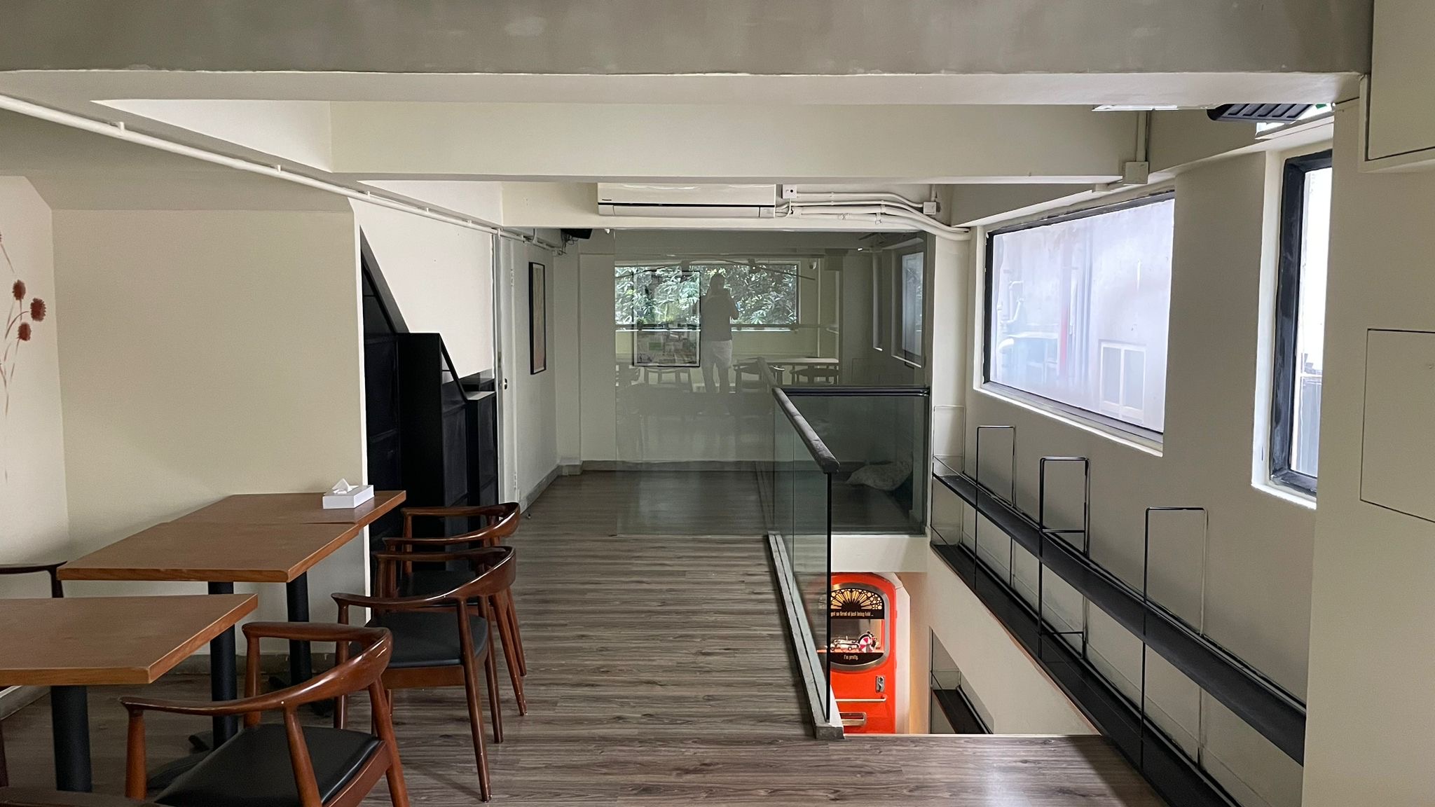 Ref#2994: A modern indoor space available to rent as restaurant, with wooden tables and chairs on the left, large windows on the right, and a glass railing overlooking an arcade game machine downstairs.