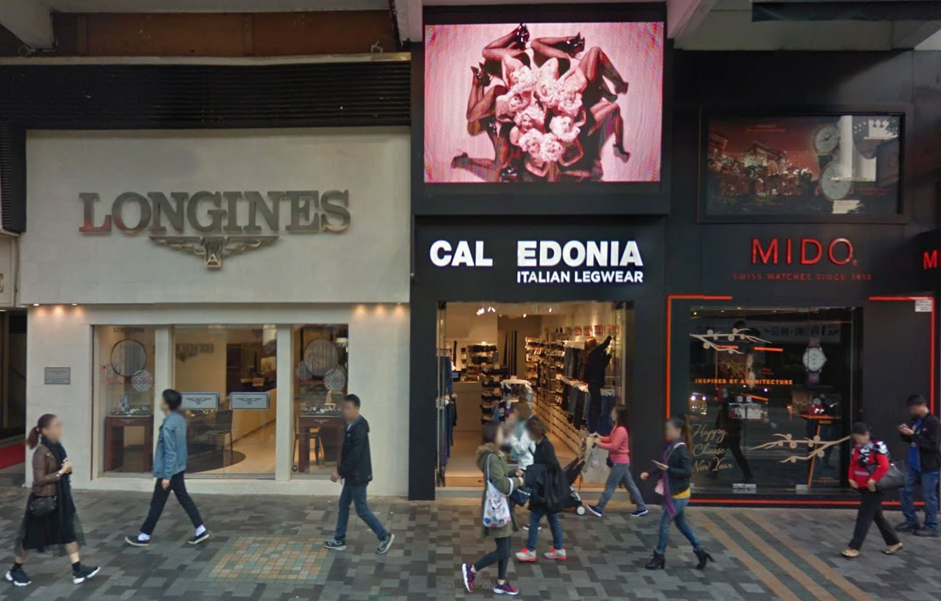People walk along Nathan Road in Tsim Sha Tsui past Longines and Mido stores under a black and white billboard.
