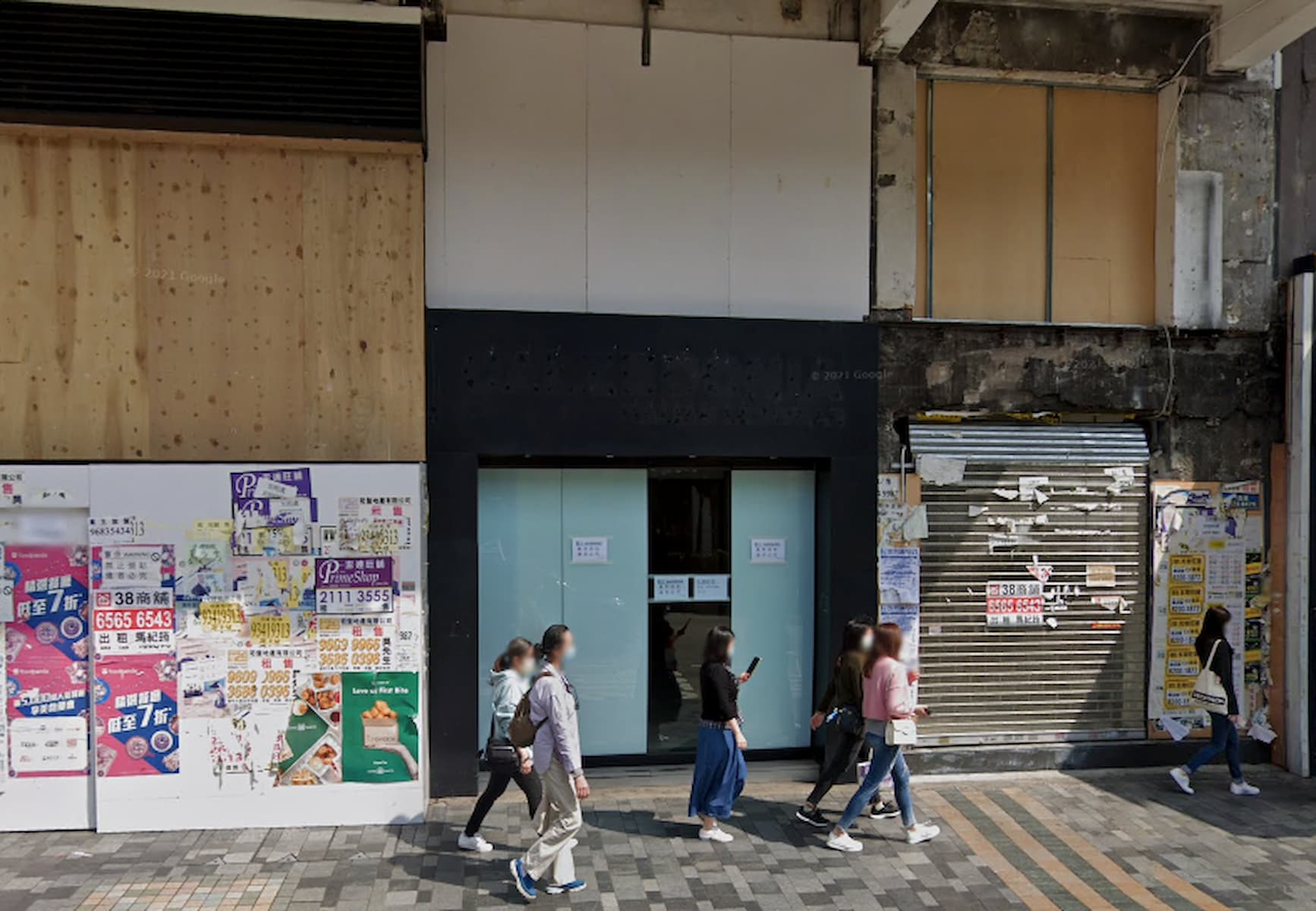 People walk by closed shops on Nathan Road with doors covered in colorful posters and signs during the day.