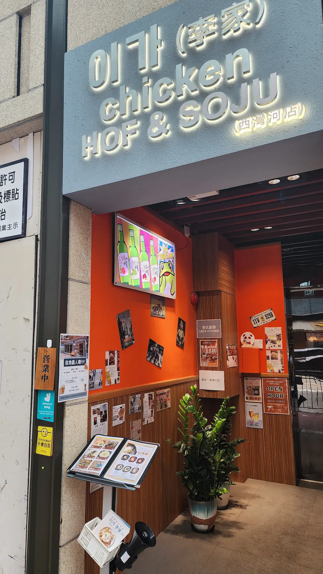 Shop on Shau Kei Wan Road, Shau Kei Wan. Chicken and soju restaurant entrance with a menu stand, wall photos, and a plant visible.