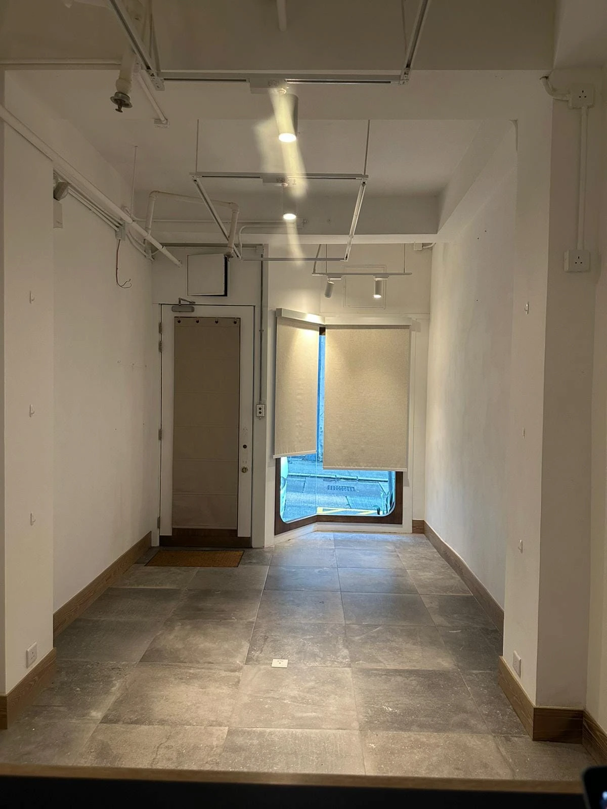 Empty room on Second Street, Sai Ying Pun, with tiled floor, white walls, ceiling pipes, a door and a window with blind.