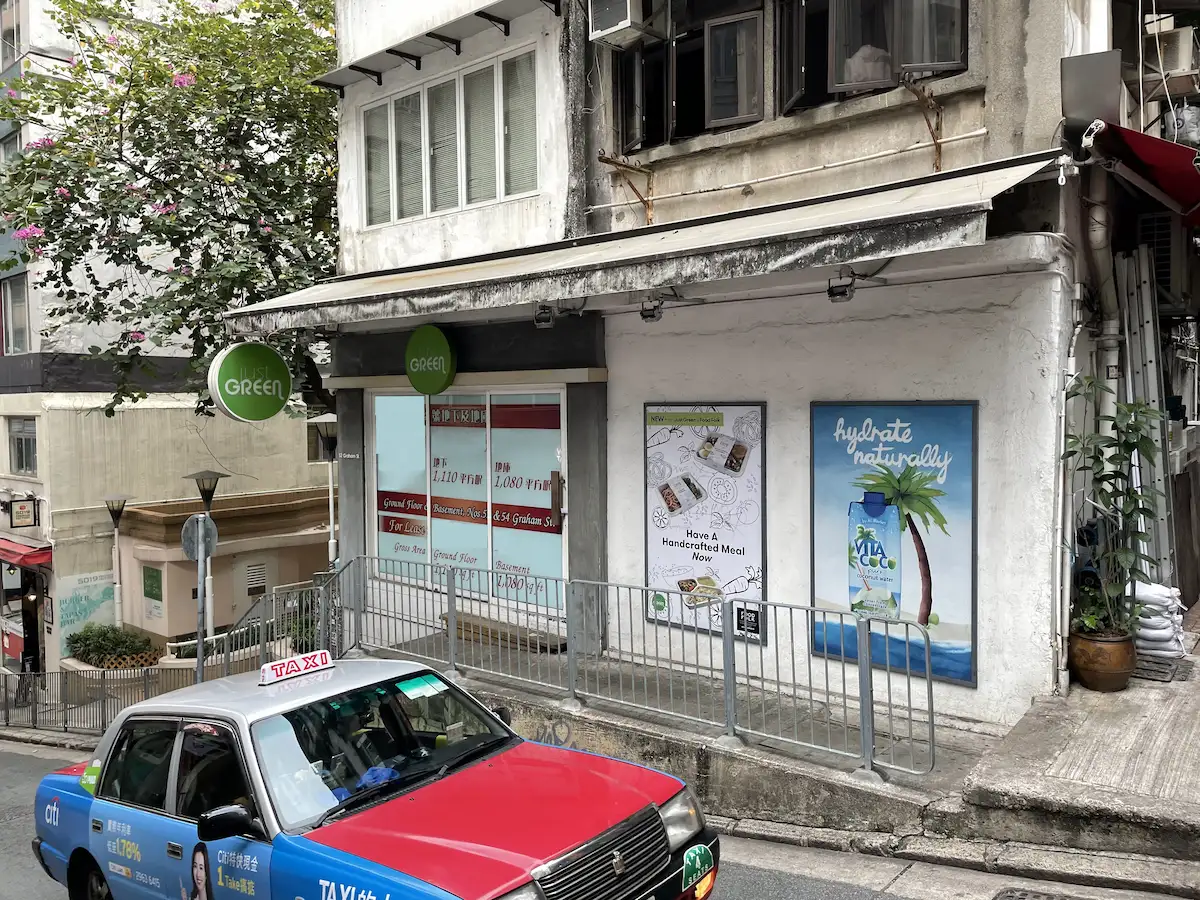 A red and blue taxi passes a storefront on sloped Graham Street in Central, with various signs and posters visible.