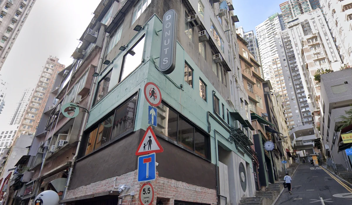 A narrow building with a DONUTS sign on Elgin Street, Central, next to taller buildings and a sloped road.