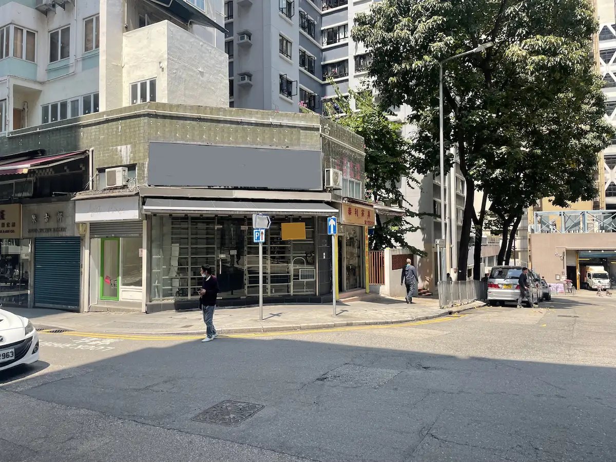A person stands on Hollywood Road in Sheung Wan beside closed shops, tall buildings, and trees on a sunny day.