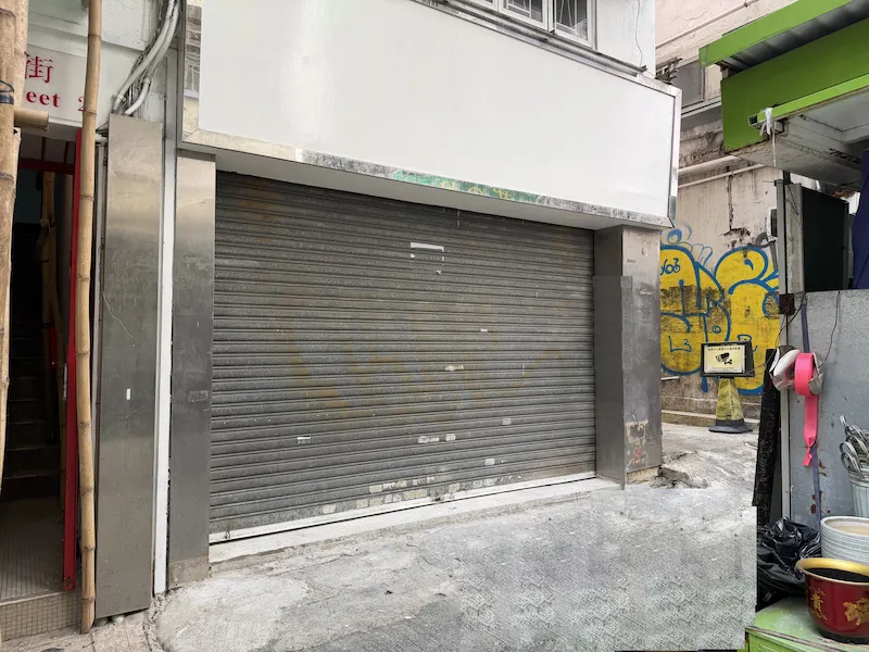 Shop on Gresson Street, Wan Chai. Closed gray metal roll-up door, with blank white sign above and graffiti nearby.