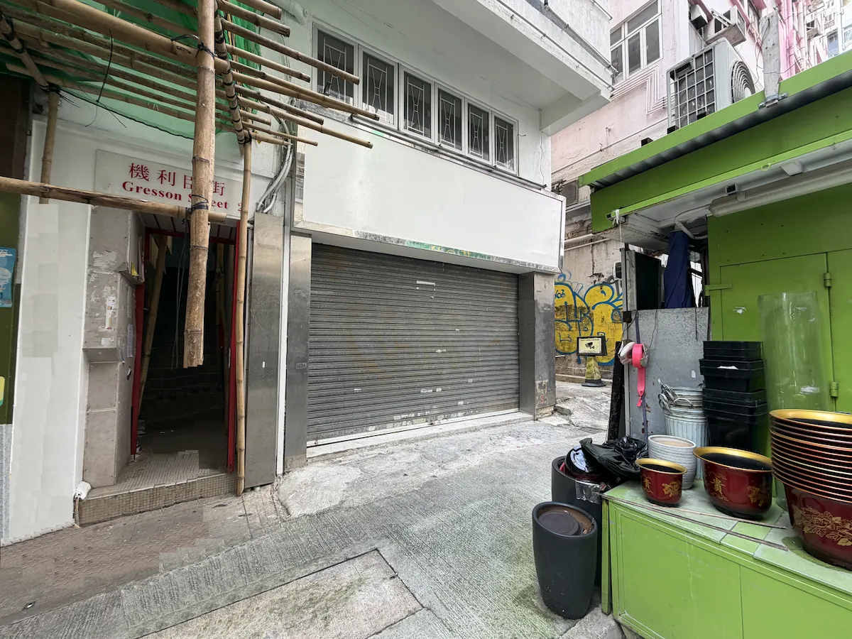 Closed metal shop shutter on Gresson Street, green food stall bowls, and alleyway in Wan Chai with bamboo scaffolding.