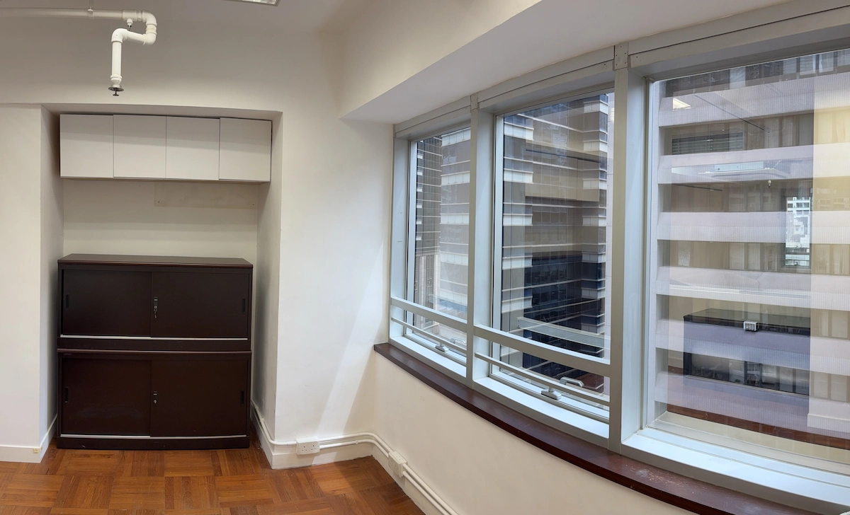 Office corner with large windows, brown cabinet, white shelves, overlooking Queen’s Road Central building exterior.