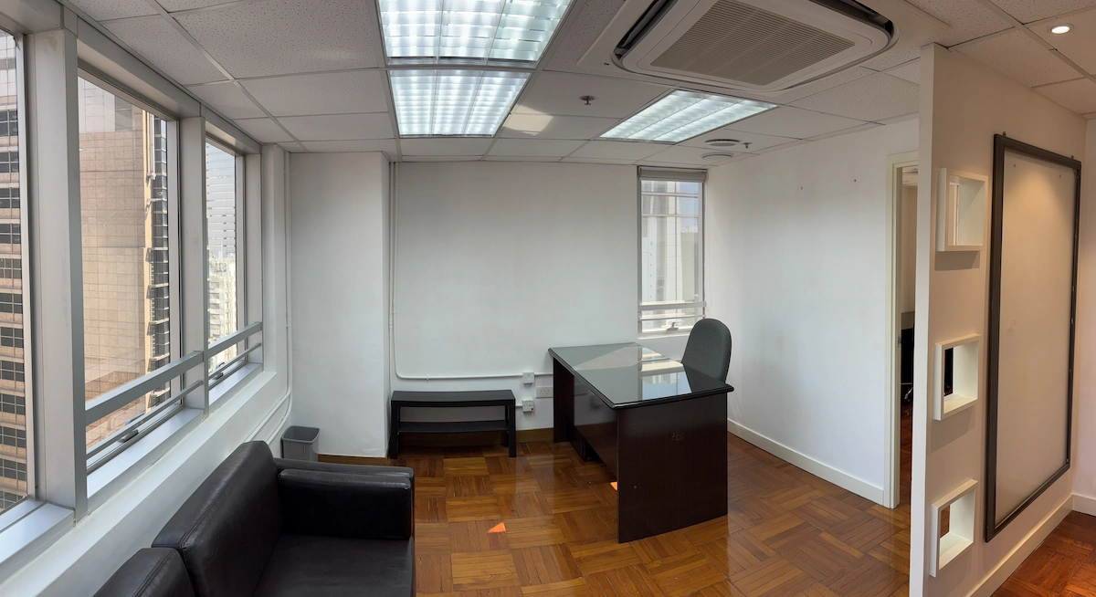 Office with desk, chair, black sofa, wood floor, white walls, and large windows overlooking Queen’s Road Central cityscape.