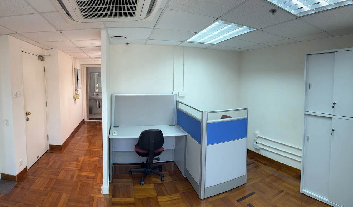 Office with two workstations, rolling chair, lockers, wood flooring, and white walls under fluorescent lighting.