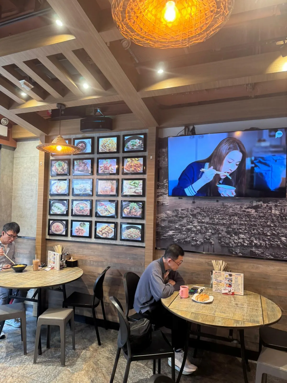 Two people sit at round tables in an Ap Lei Chau Main Street restaurant with food photos and a TV showing a woman eating.