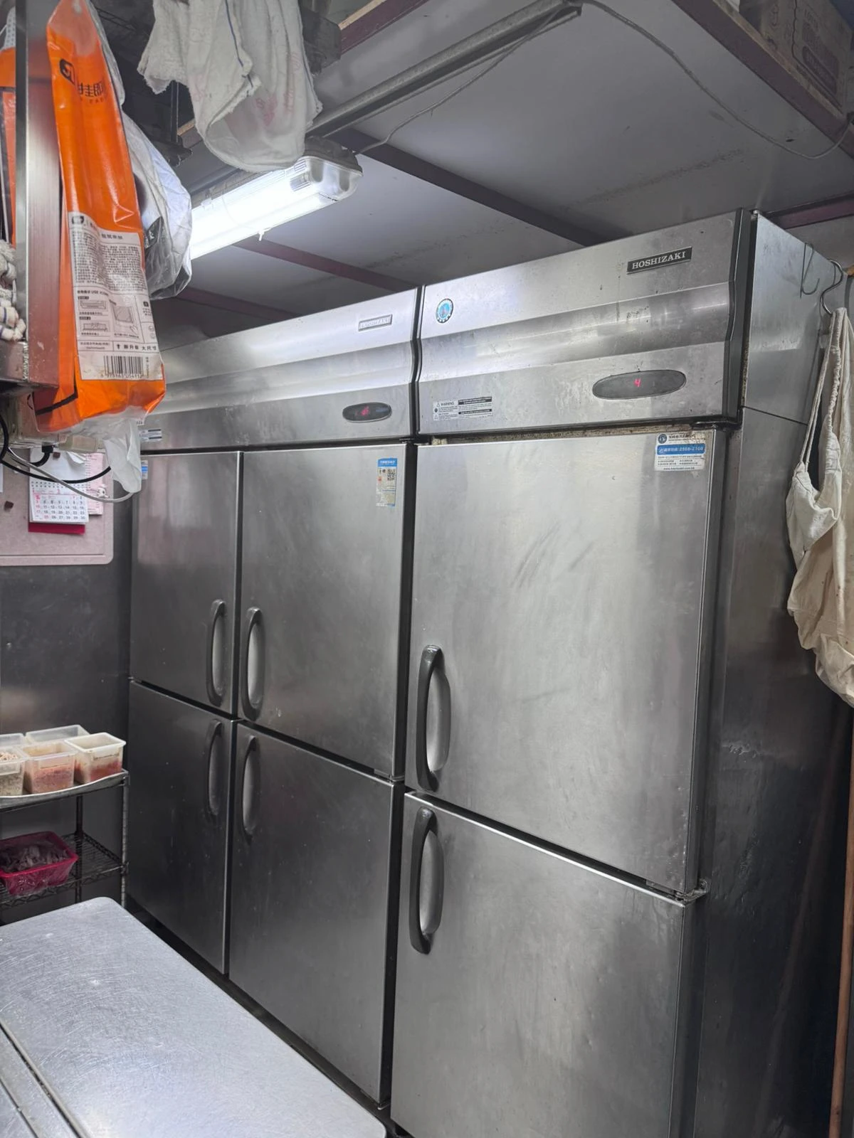 Four stainless steel refrigerators are lined up in an Ap Lei Chau Main Street kitchen with shelves and containers nearby.