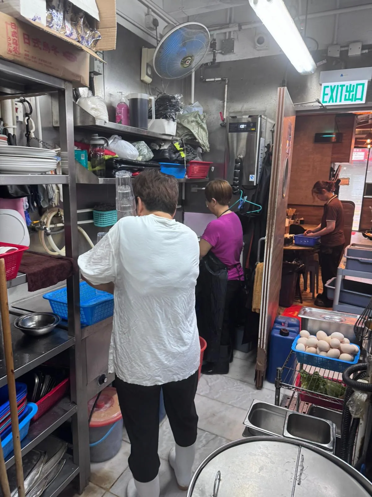 Three people work in a narrow Ap Lei Chau Main Street kitchen, washing dishes and preparing food near shelves and supplies.