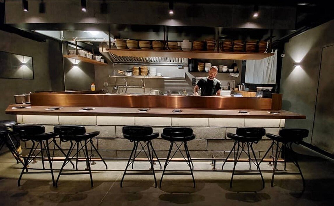 A chef stands behind a modern bar counter in a dimly lit restaurant on Pottinger Street in Central with empty stools.