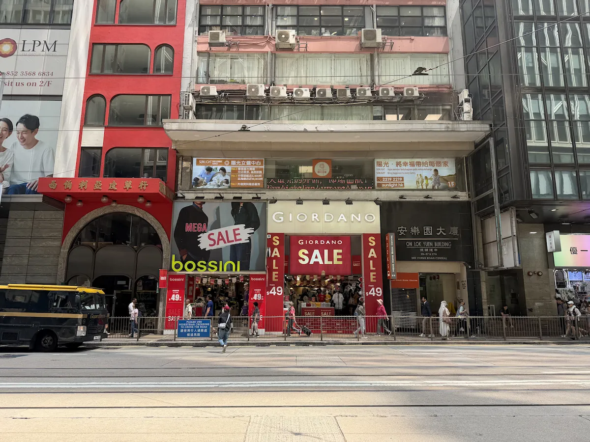 Street view of shops on Des Voeux Road Central with Giordano’s red signs visible and pedestrians walking past.