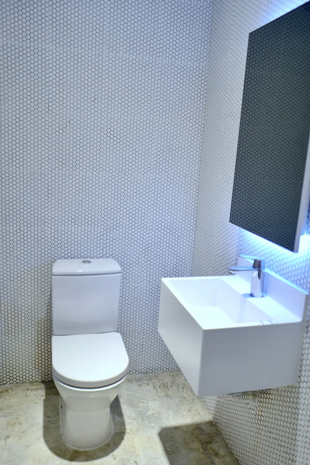 Bathroom in Sheung Wan featuring a white toilet, wall-mounted sink, illuminated mirror, and hexagonal wall tiles.