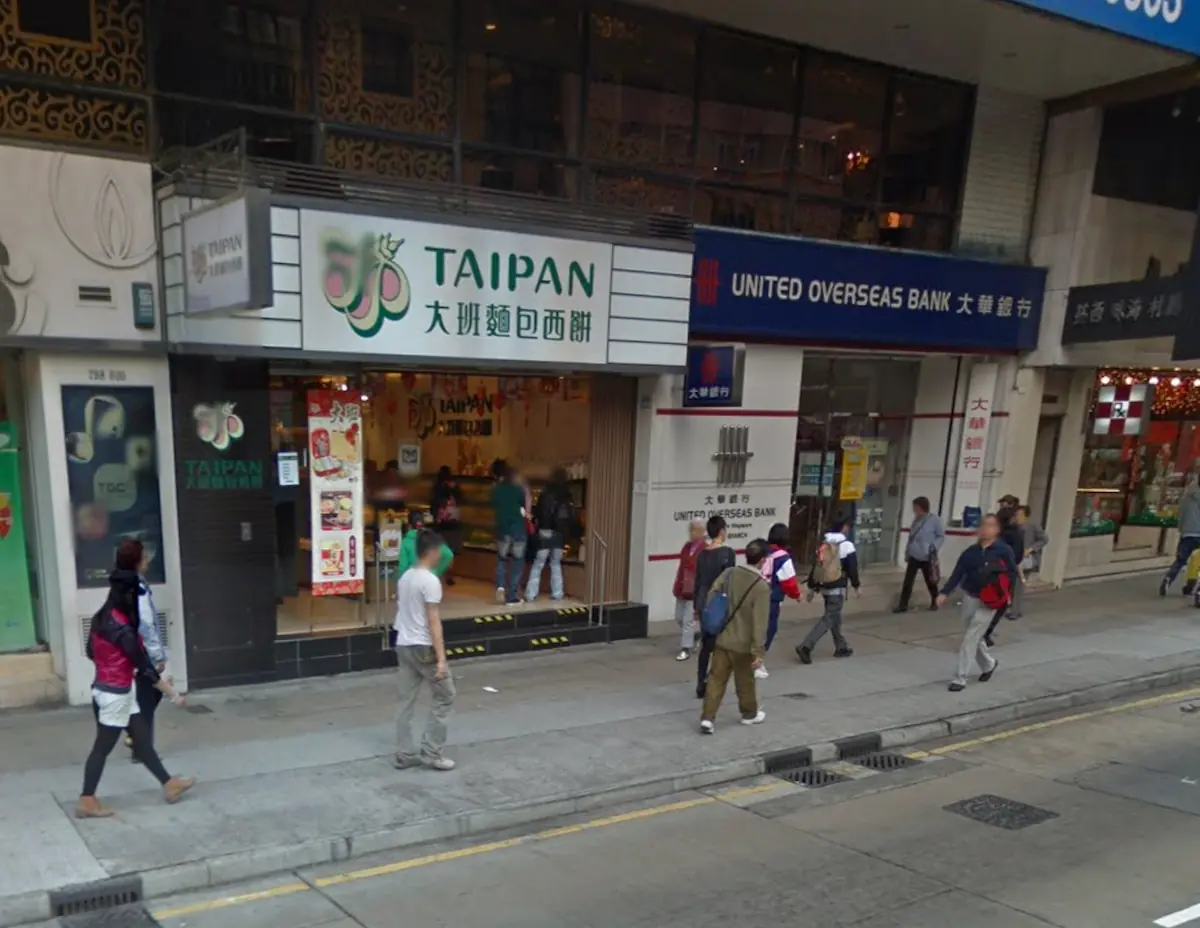 Pedestrians walk by Taipan bakery and United Overseas Bank on Nathan Road in Mong Kok with shops and signs visible.
