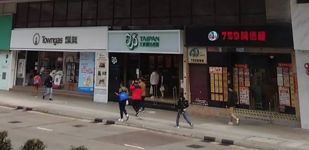 Four people walk on the Nathan Road sidewalk in Mong Kok, passing Towngas, Taipan, and a store with red signage.