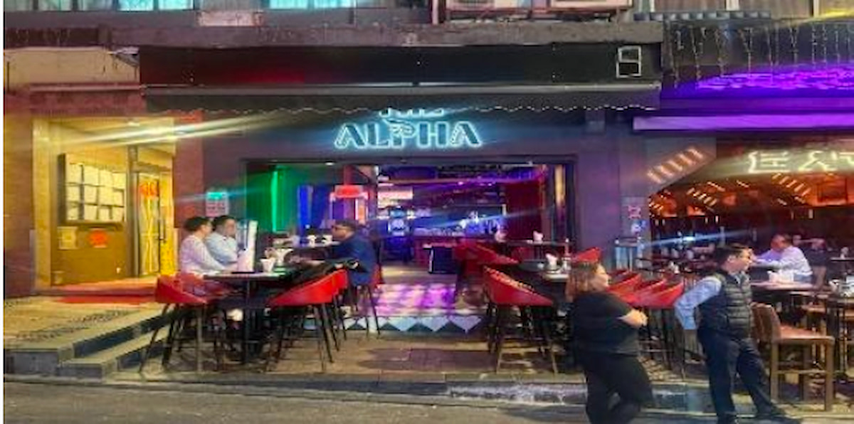 People are seated at outdoor tables on Wing Wah Lane in Central outside ALPHA bar under colorful lights at night.