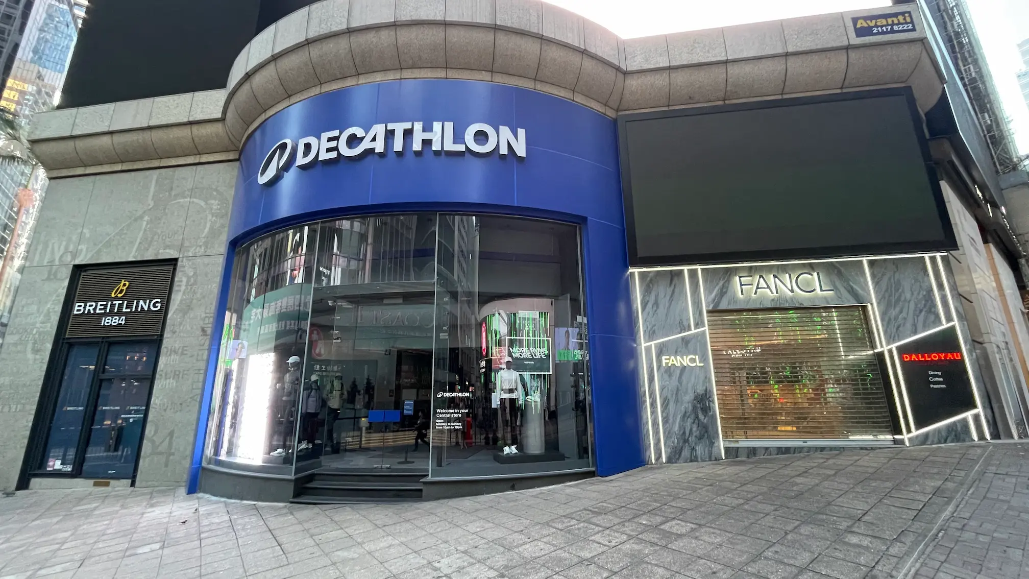 Curved building on Queen’s Road Central displaying Decathlon, Breitling, and FANCL shops with glass window displays.
