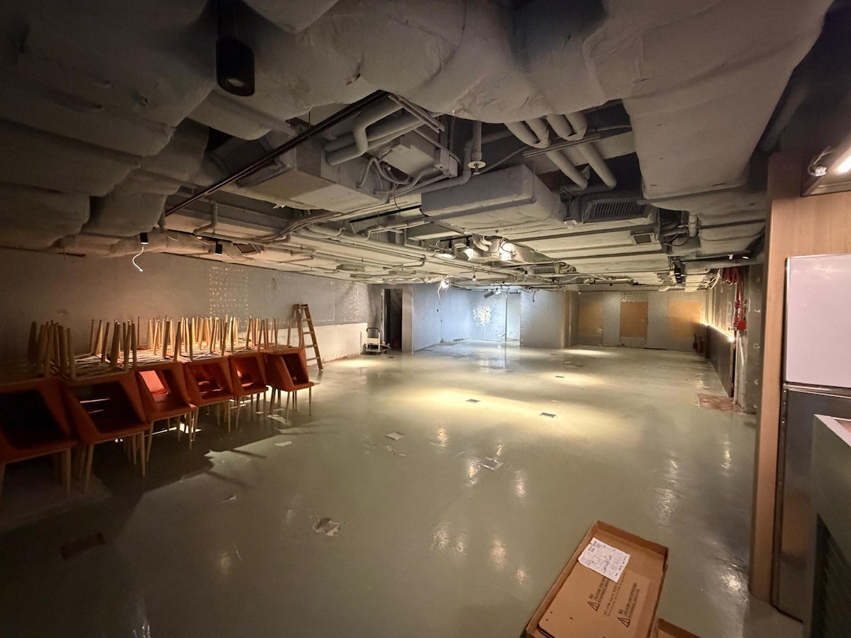 Large empty room on Wellington Street with exposed pipes, stacked chairs on the left, and boxes in the foreground.