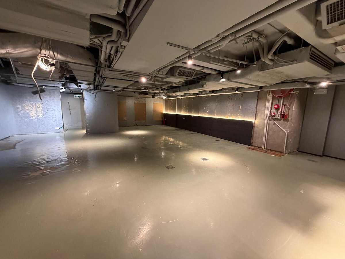 Empty industrial room on Wellington Street with concrete floor, exposed ceiling pipes, wall lights, and fire equipment.