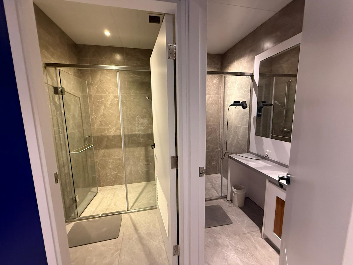 Two modern bathrooms at Yip Kan Street, Wong Chuk Hang with glass showers, beige tiles, sink, mirror and hairdryer.
