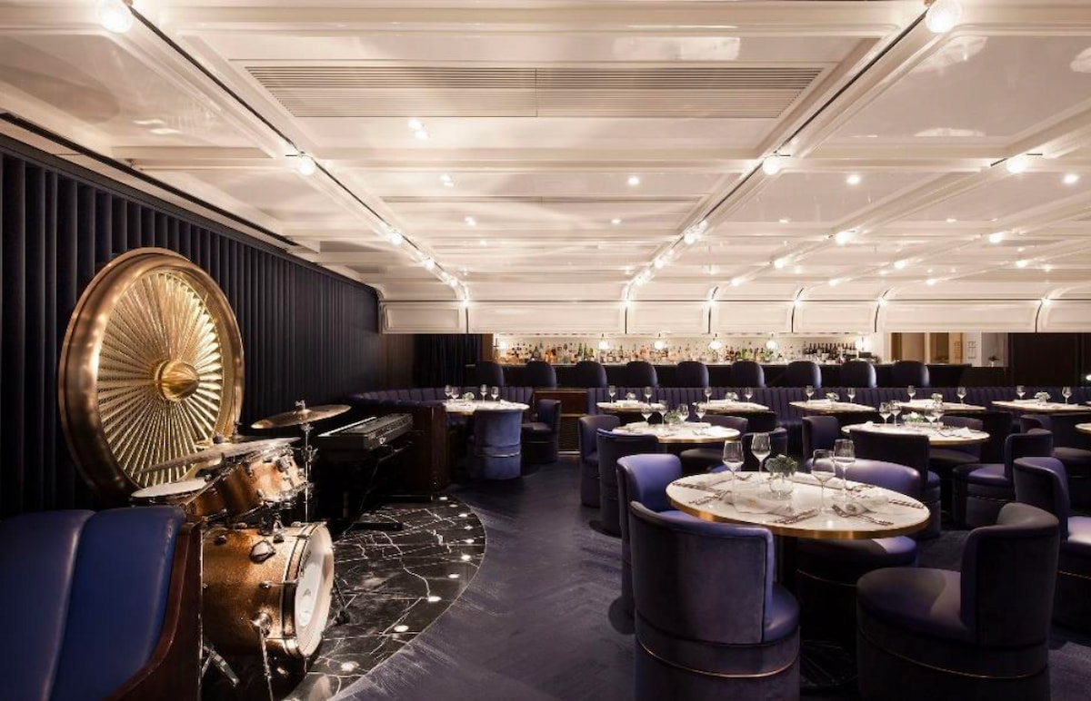 Central restaurant with round tables, blue chairs, a drum set, piano, and a large Duddell Street wall artwork.