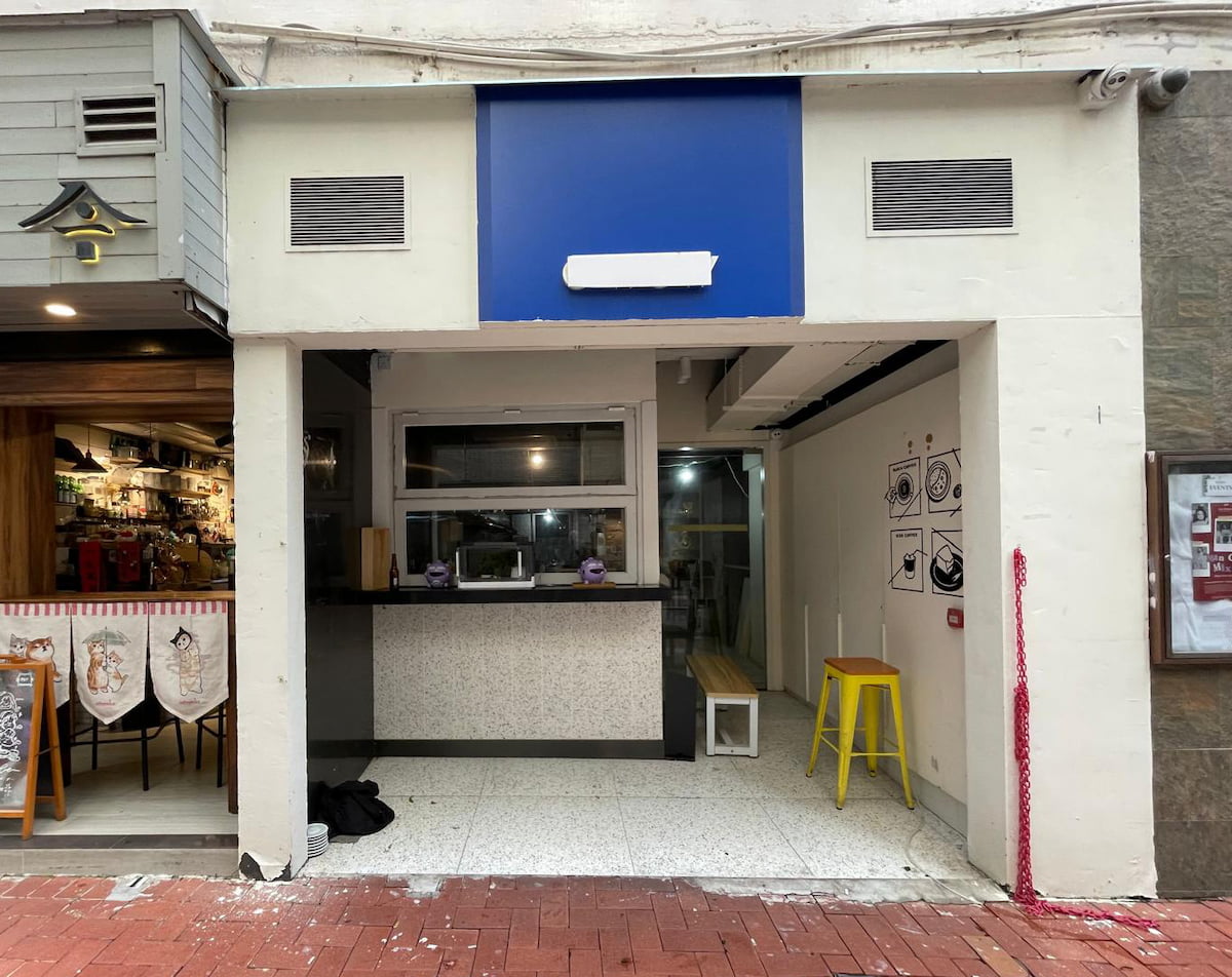 Storefront on Hillier Street in Sheung Wan with a blank blue sign, yellow stool, wall art, and open entrance.