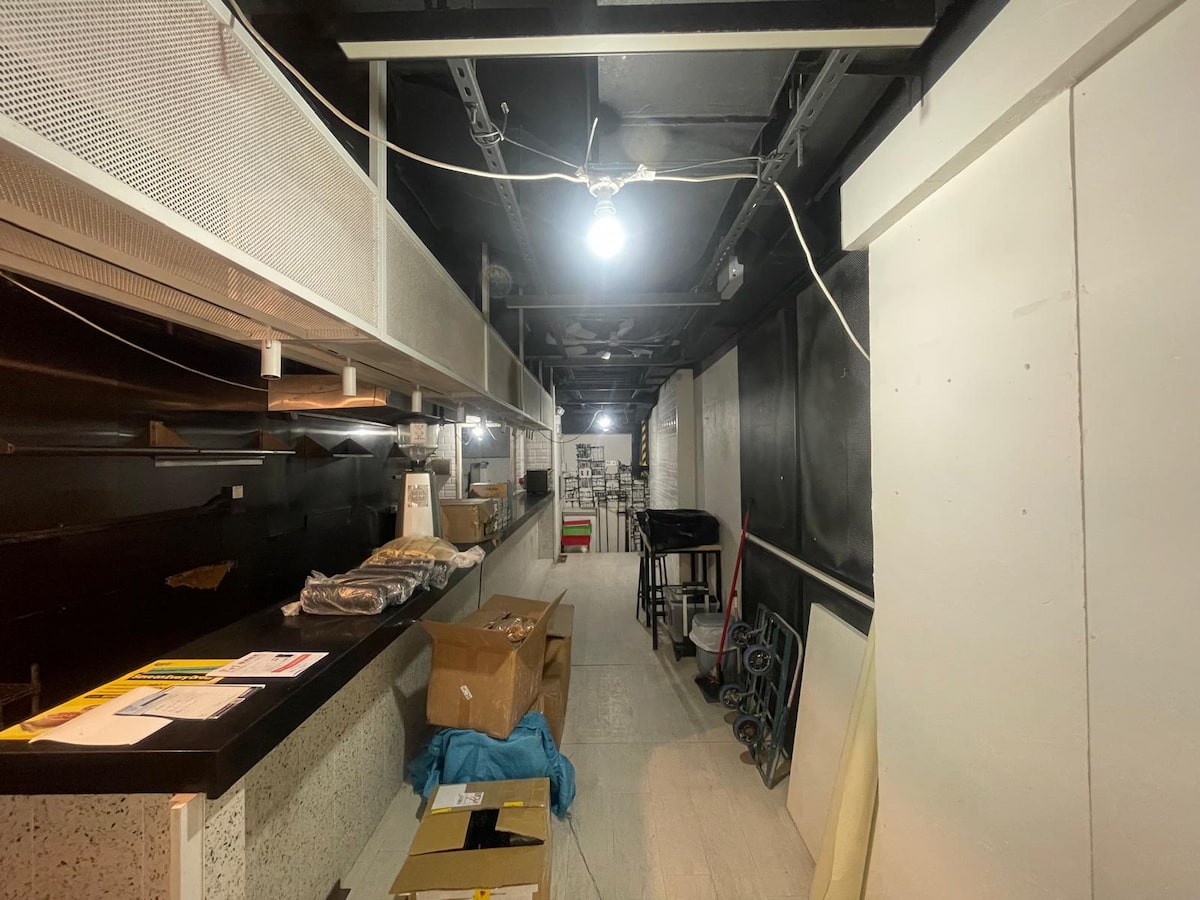 Narrow Hillier Street room with counter on the left, boxes on floor, stacked furniture behind, lit by bare bulb.