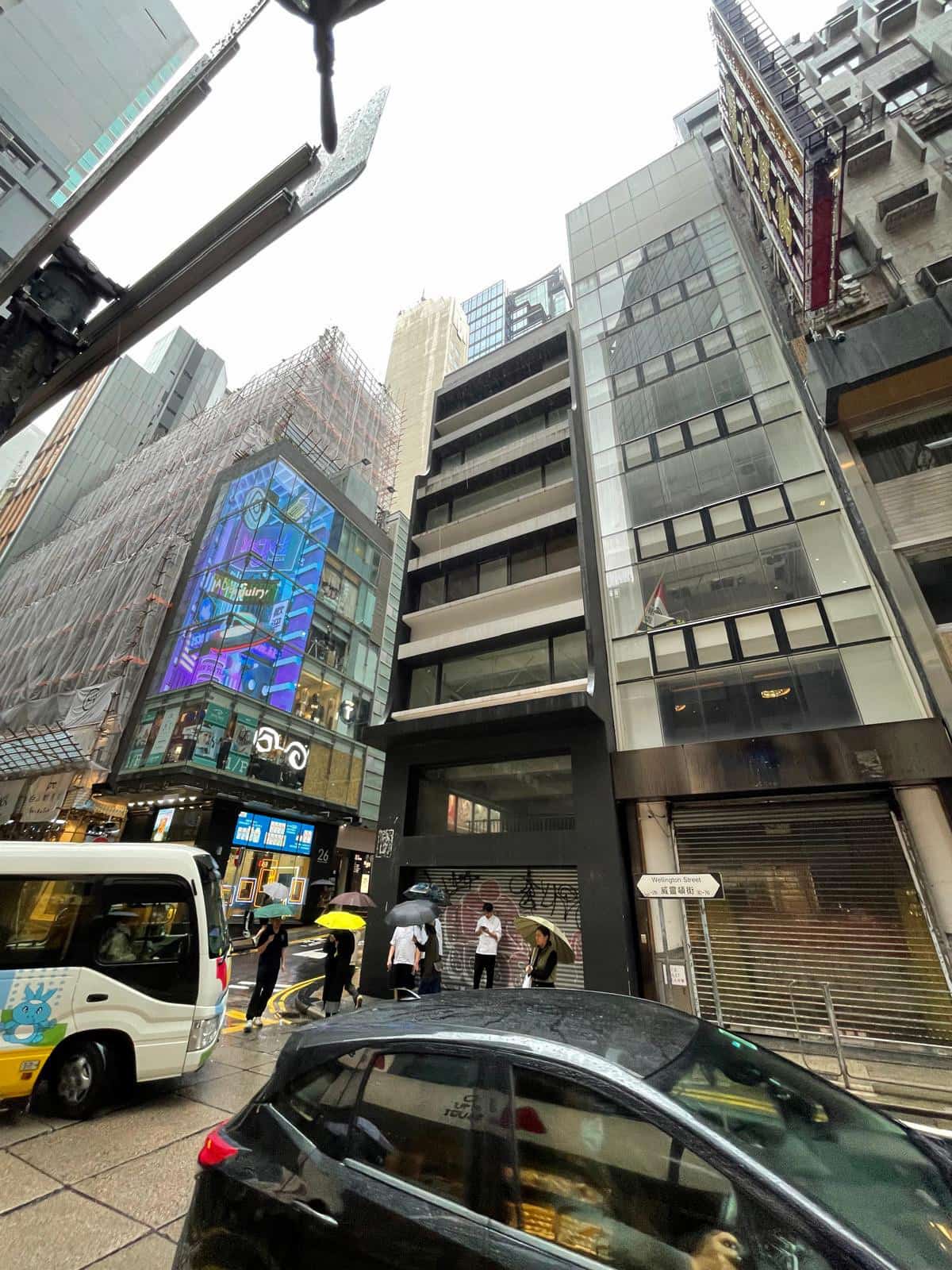 People with umbrellas walk on rainy Wellington Street in Central among tall buildings, cars, and a digital billboard.