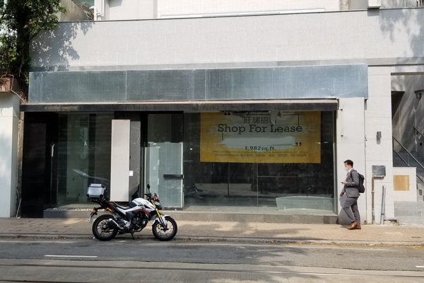 A man walks past an empty storefront with a sign on Wong Nai Chung Road, motorcycle parked in front.