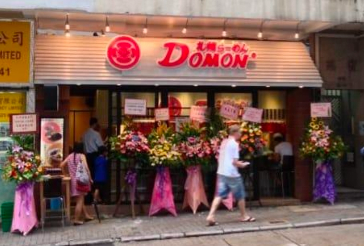 People walk by a ramen restaurant with floral stands and signs on Caine Road in Mid-Levels, a busy street.