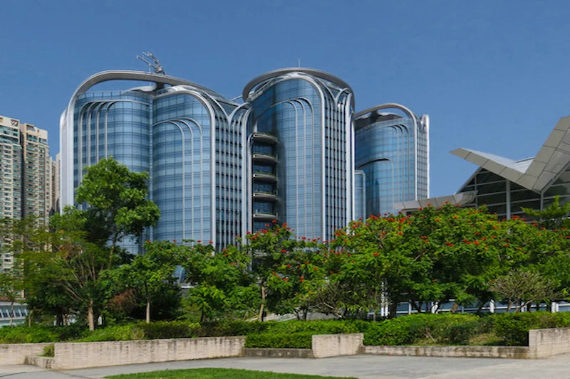 Modern glass building with curved facades in West Kowloon, trees in front and clear blue sky in the background.