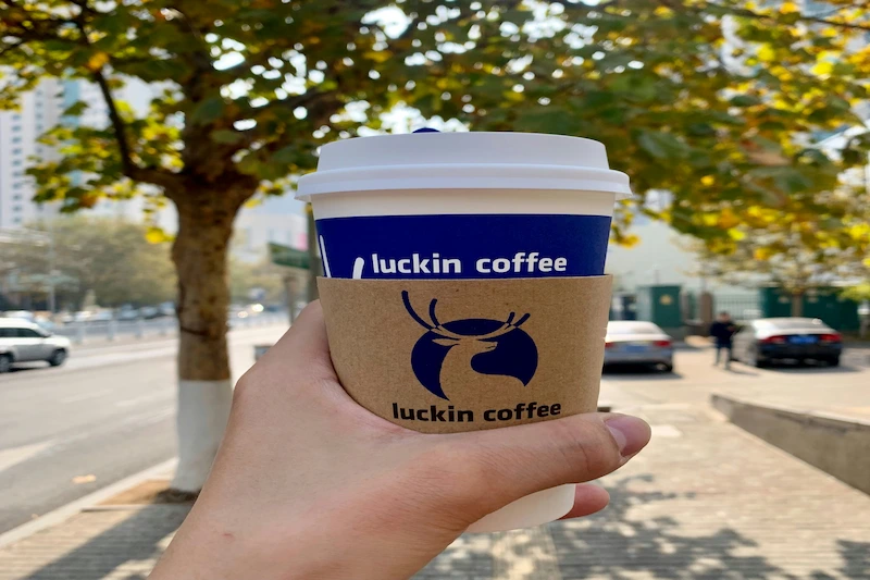 A hand holds a Luckin Coffee cup on a sunny Hong Kong street lined with trees, cars, and tall city buildings.