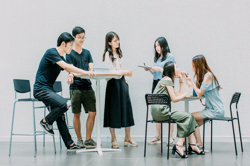 Six people socialize in a bright modern room standing and sitting around two small tables in an urban hostel.