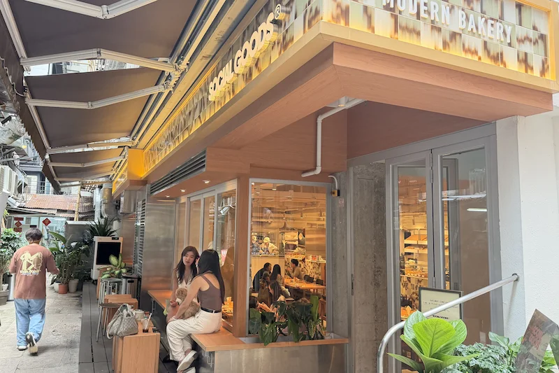 People sit outside a modern Hong Kong bakery with yellow signage; egg tarts and potted plants are near the seating area.